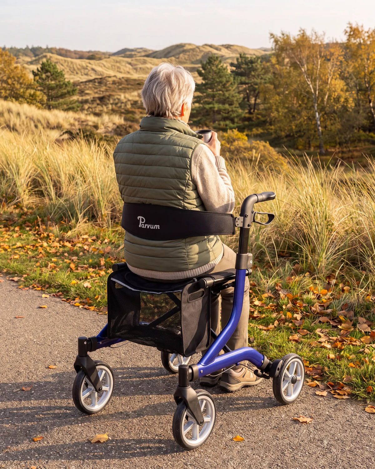 Parvum SuperLight Plus in blue beside a path; a woman sits on the rollator holding a cup of tea.